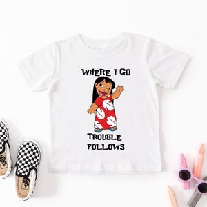 A white t-shirt with a cartoon character and text printed on it, along with a pair of black and white checkered shoes and some colored pencils.