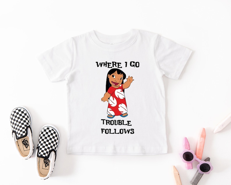 A white t-shirt with a cartoon character and text printed on it, along with a pair of black and white checkered shoes and some colored pencils.
