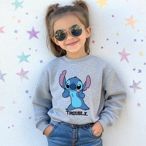 A young girl wearing a gray sweatshirt with a cartoon character and the word "TROUBLE" printed on it, smiling and posing against a starry background.