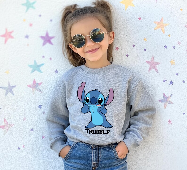 A young girl wearing a gray sweatshirt with a cartoon character and the word "TROUBLE" printed on it, smiling and posing against a starry background.