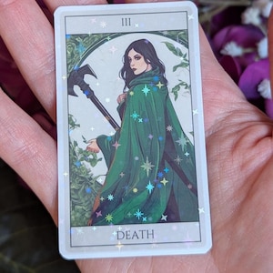 May include: A tarot card with a holographic finish. The card is white with a green border and features a woman in a green cloak holding a scythe. The card number, "III", and the word "DEATH" are printed on the card.