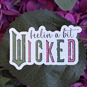 May include: A white sticker with the text "feelin' a bit WICKED" in green, pink, and white letters with glitter accents. The sticker is on a green leaf with purple flowers in the background.