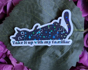 Könnte beinhalten: Ein weißer Aufkleber mit einer schwarzen Katzensilhouette auf einem violetten Blumenhintergrund. Die Katze ist mit schimmerndem Glitzer bedeckt und der Aufkleber sagt "Take it up with my familiar".