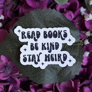 May include: White sticker with black text that reads "Read Books Be Kind Stay Weird" with glitter accents. The sticker is on a green leaf with purple flowers.