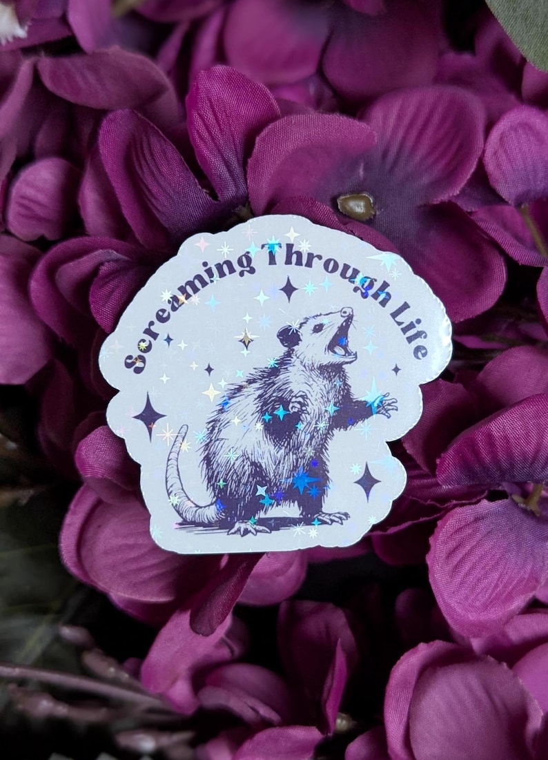May include: A white sticker with a holographic finish features a black and white illustration of a possum with its mouth open, surrounded by stars. The text "Screaming Through Life" is written in a retro font around the possum.
