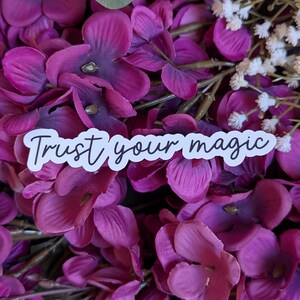 May include: White sticker with the text "Trust your magic" in black cursive lettering, placed on a background of pink and white flowers.