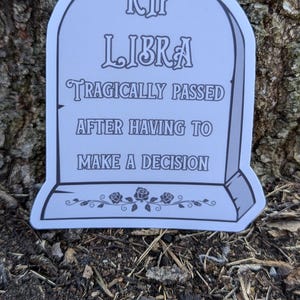 May include: A white tombstone with the text "RIP LIBRA TRAGICALLY PASSED AFTER HAVING TO MAKE A DECISION" and a floral design at the bottom.