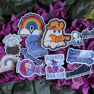 May include: A collection of colourful, glittery stickers with various witchy and spooky themes. The stickers feature phrases like "Let's Hex the Patriarchy Together", "Cast Spells, Not Shade", "In My Witchy Aunt Era", "Salem Witch Book Club", "Sorry My Horoscope Says I Can't Go", and "Take It Up With My Familiar".