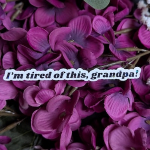 May include: A sticker with the text "I'm tired of this, grandpa!" on a background of purple flowers.