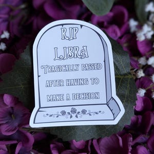 May include: A white sticker with a tombstone design that reads "RIP LIBRA tragically passed after having to make a decision". The sticker is on a background of purple flowers.