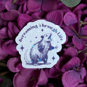 May include: A white sticker with a holographic finish featuring a black and white illustration of an opossum with its mouth open, surrounded by stars. The text "Screaming through Life" is written in a curved font around the opossum.