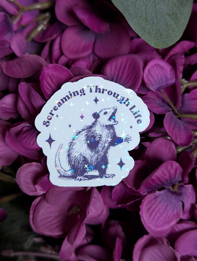 May include: A white sticker with a holographic finish featuring a black and white illustration of an opossum with its mouth open, surrounded by stars. The text "Screaming through Life" is written in a curved font around the opossum.