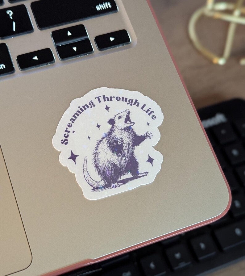 May include: A white sticker with a black and white illustration of a possum screaming with stars around it. The text "Screaming Through Life" is written in a retro font around the possum.