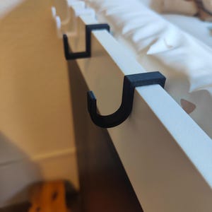 Könnte beinhalten: Drei schwarze Plastikhaken, die an einer weißen Oberfläche befestigt sind. Die Haken sind J-förmig und zum Aufhängen von Gegenständen gedacht.