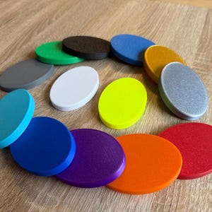 May include: A collection of 12 oval-shaped coasters in various colours, including teal, blue, purple, orange, red, yellow, white, grey, and black. The coasters are arranged in a circle on a wooden surface.