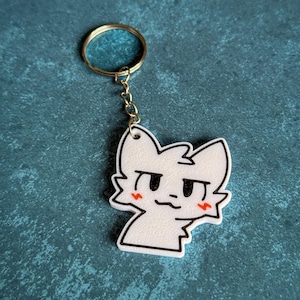 May include: White keychain featuring a cartoon cat face with black eyes and a red blush. The cat is looking to the right. The keychain is attached to a gold key ring.
