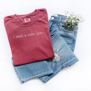 May include: A red t-shirt with the text "i need a coke zero."  The t-shirt is folded on top of a pair of blue jeans.