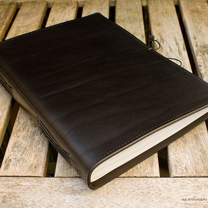 May include: A black leather-bound journal with a stitched spine and a leather strap closure. The journal is lying open on a wooden surface.