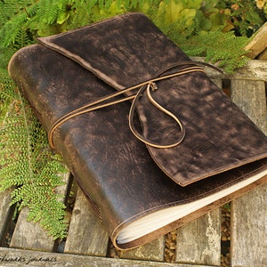 May include: A dark brown leather journal with a wrap-around flap and a leather tie closure. The journal is open, revealing white pages. The cover has a distressed, textured appearance. The journal is resting on a wooden surface.