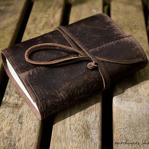 May include: A brown leather-bound journal with a leather strap closure. The journal is closed and lying on a wooden surface.