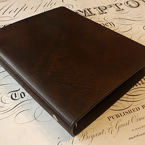 May include: A brown leather binder with a ring mechanism. The binder is closed and lying on a white surface with black calligraphy.