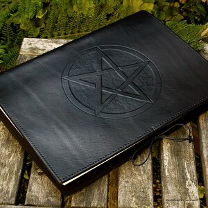 May include: A black leather-bound journal with a pentagram embossed on the front cover. The journal is closed and lying on a wooden surface.