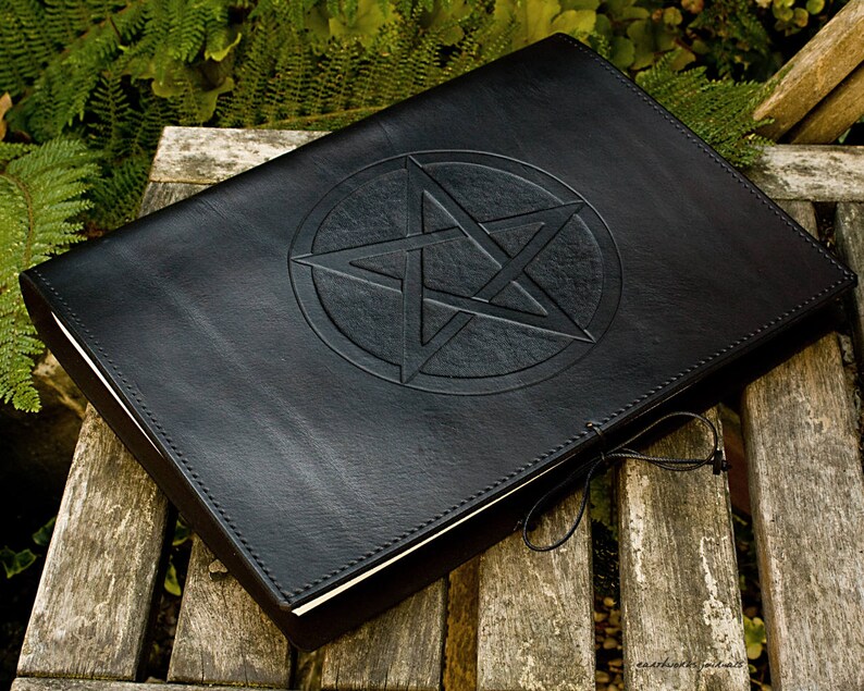 May include: A black leather-bound journal with a pentagram embossed on the front cover. The journal is closed and lying on a wooden surface.