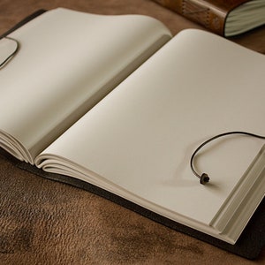 May include: A brown leather-bound journal with blank pages and black leather ties. The journal is open and lying flat on a brown surface.