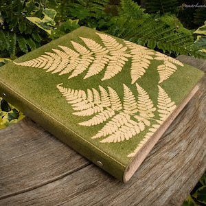 May include: A green journal with a fern design. The cover is a textured green with two large, light-colored fern leaf imprints. The journal is resting on a weathered wooden surface, with greenery in the background. The text "earthworks journals" is in the top right corner.