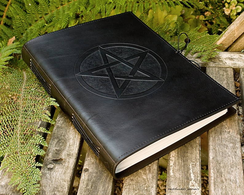 May include: A black leather-bound journal with a pentagram embossed on the front cover. The journal has a leather strap closure and is lying on a wooden surface.