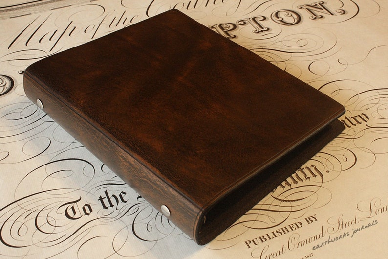 May include: A dark brown leather-bound journal or binder with silver rivets. The cover has a textured appearance. The background features a vintage map design with elegant calligraphy.
