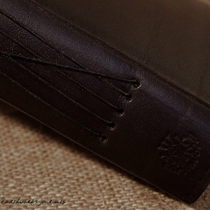 May include: Close-up of a dark brown leather-bound journal with a stitched spine and a small logo on the cover.