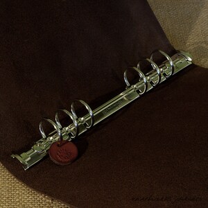 May include: A silver-colored binder mechanism with six rings, resting on a dark brown, textured surface. A small, round, reddish-brown leather tag hangs from the mechanism. The text "DENSONS @ BIRMINGHAM" is visible on the metal.