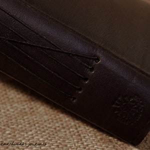 May include: Close-up of a dark brown leather-bound journal with a stitched spine and a small embossed logo on the cover.