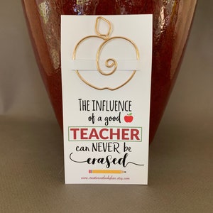 May include: Gold wire apple bookmark with the text "The influence of a good teacher can never be erased" and the website www.creativeoutlookglass.etsy.com.