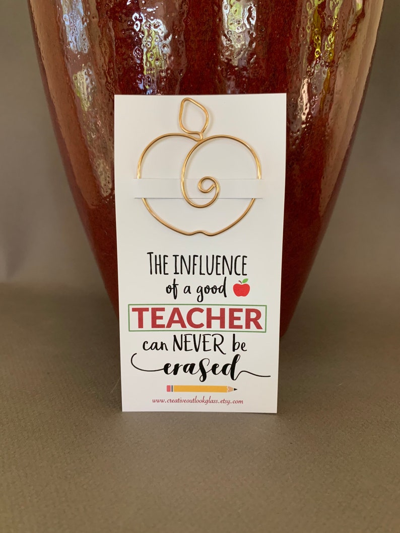 May include: Gold wire apple bookmark with the text "The influence of a good teacher can never be erased" and the website www.creativeoutlookglass.etsy.com.