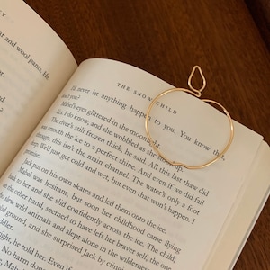 Puede incluir: Un marcap&aacute;ginas de oro en forma de manzana con un acabado liso y brillante. El marcap&aacute;ginas est&aacute; colocado en un libro abierto con texto sobre un r&iacute;o helado. El texto dice "The Snow Child. I'll never let anything happen to you. You know that, don't you?"