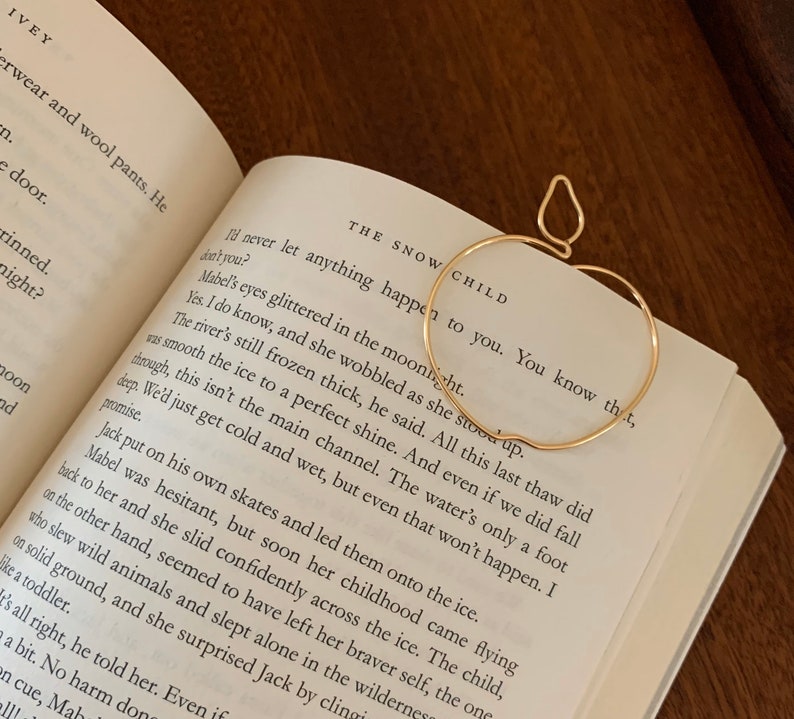 Puede incluir: Un marcap&aacute;ginas de oro en forma de manzana con un acabado liso y brillante. El marcap&aacute;ginas est&aacute; colocado en un libro abierto con texto sobre un r&iacute;o helado. El texto dice "The Snow Child. I'll never let anything happen to you. You know that, don't you?"