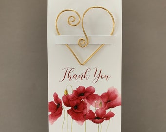 Puede incluir: Marcador de libros en forma de corazón de alambre dorado con una tarjeta blanca que dice "Gracias" y tiene un diseño de acuarela de amapolas rojas. La tarjeta también incluye la dirección del sitio web "www.creativeoutlookglass.etsy.com".