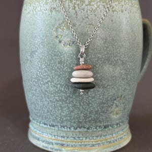 Puede incluir: Collar de cadena plateada con un colgante de piedras lisas apiladas. Las piedras son en tonos marrón, blanco y verde. El collar se muestra frente a una taza de cerámica verde claro.