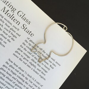 May include: A silver metal rod shaped like a teardrop with a rounded end. The rod is on a white piece of paper with text about heating glass to a molten state.