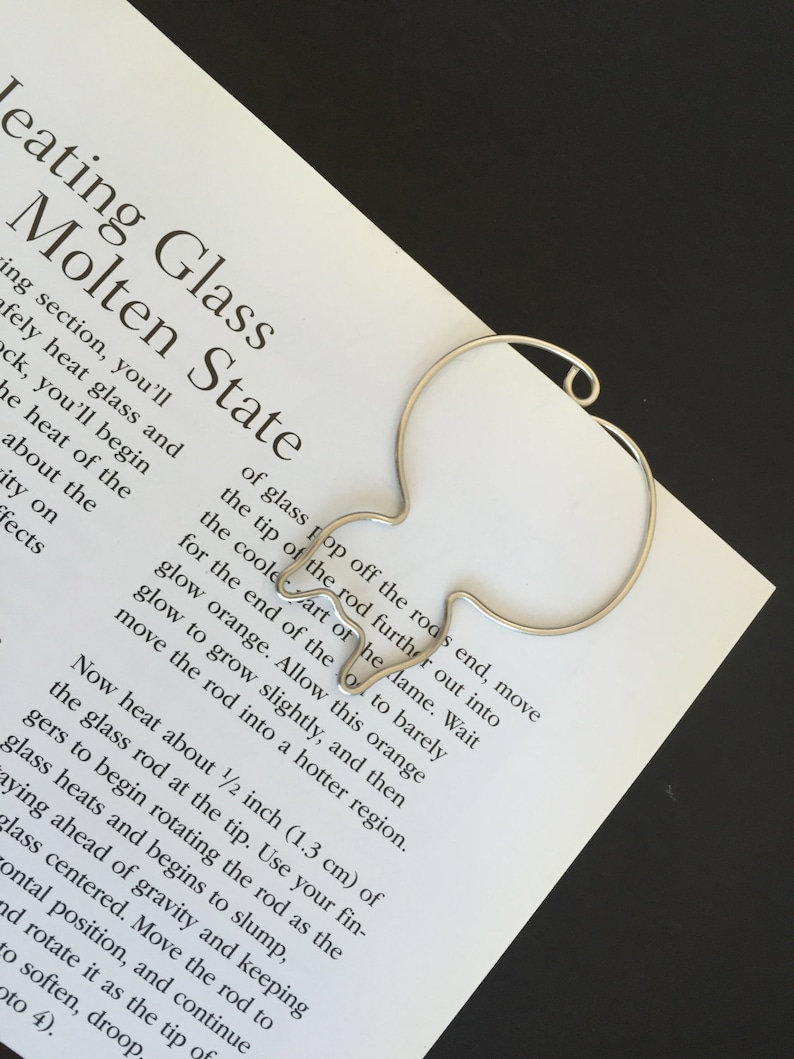 May include: A silver metal rod shaped like a teardrop with a rounded end. The rod is on a white piece of paper with text about heating glass to a molten state.