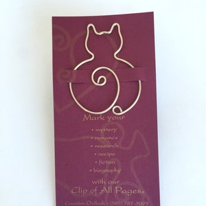 May include: A silver wire bookmark shaped like a cat with a curled tail. The bookmark is on a burgundy card with gold text that reads "Mark your... with our Clip of All Pages&reg;".