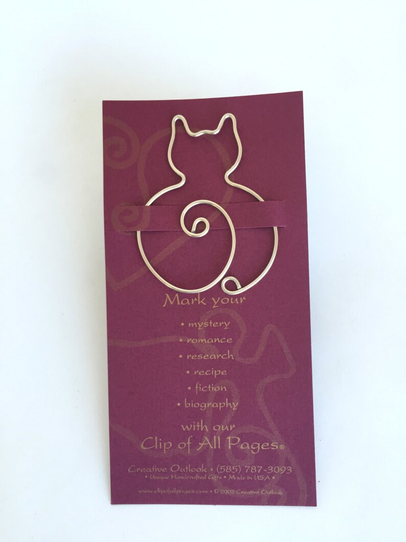 May include: A silver wire bookmark shaped like a cat with a curled tail. The bookmark is on a burgundy card with gold text that reads "Mark your... with our Clip of All Pages&reg;".