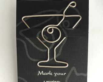 May include: Silver wire martini glass bookmark with an olive. The bookmark is on a black background with the text "Mark your" and a list of words: mystery, romance, research.