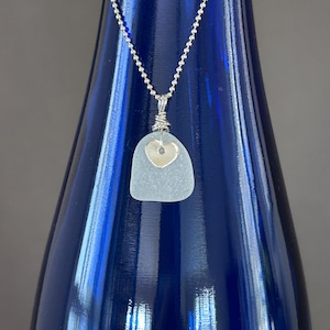 May include: A silver chain necklace with a pendant made of sea glass. The pendant is a light blue, irregular shape with a small, silver heart in the center. The necklace is displayed against a dark blue background.