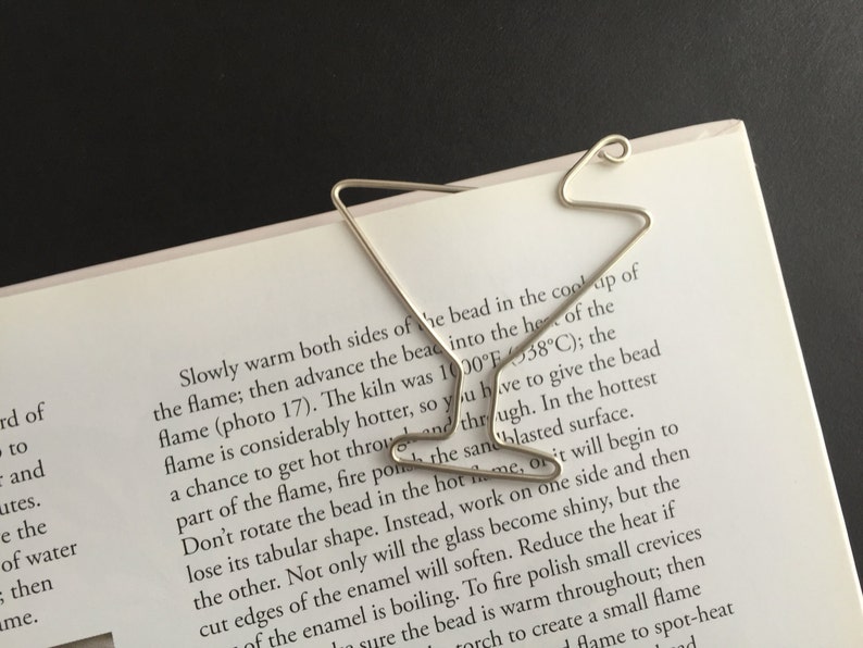 K&ouml;nnte beinhalten: Ein silbernes Lesezeichen in Form eines Martini-Glases. Das Lesezeichen liegt auf einer Seite eines Buches mit Text &uuml;ber Glasblasen.