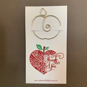 Puede incluir: Una tarjeta blanca con un dise&ntilde;o de manzana de alambre dorado y el texto "Gracias" en un dise&ntilde;o de manzana en forma de coraz&oacute;n rojo. La parte inferior de la tarjeta dice "www.creativeoutlookglass.etsy.com".