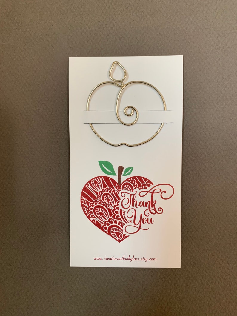 Puede incluir: Una tarjeta blanca con un dise&ntilde;o de manzana de alambre dorado y el texto "Gracias" en un dise&ntilde;o de manzana en forma de coraz&oacute;n rojo. La parte inferior de la tarjeta dice "www.creativeoutlookglass.etsy.com".