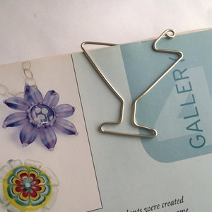 K&ouml;nnte beinhalten: Ein silbernes Lesezeichen aus Metall in Form eines Martini-Glases. Das Lesezeichen liegt auf einem Buch mit dem Wort "GALLERY" auf dem Einband.